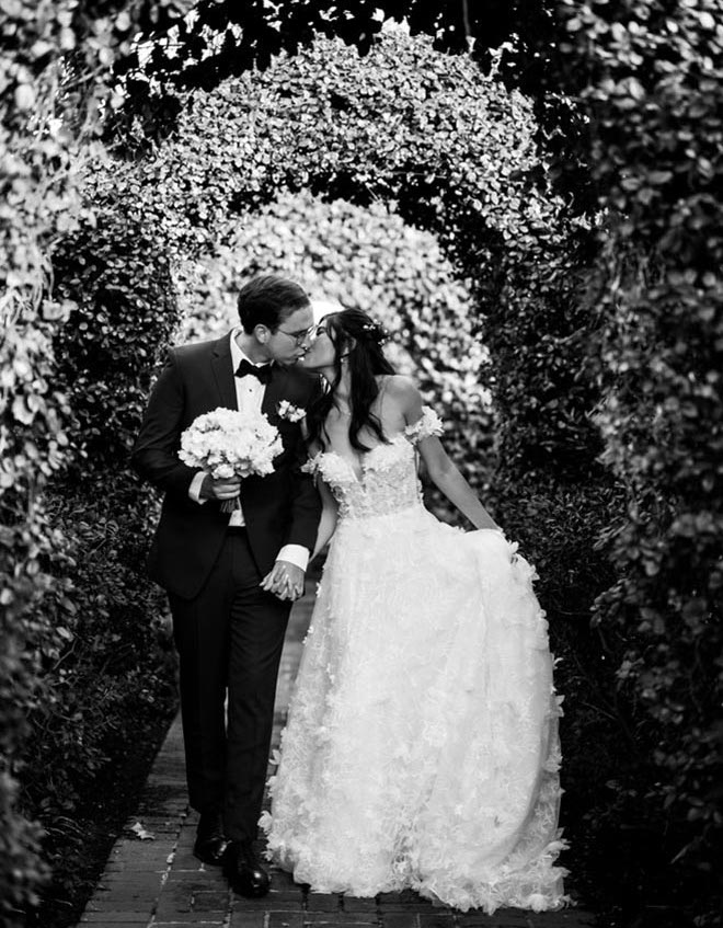 The bride and groom kiss under a archway of greenery while holding hands.