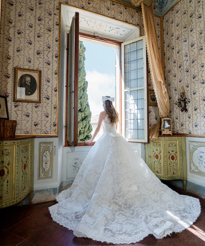 Model looking out a window wearing a bow in her hair and an all lace gown with bows down the back. 
