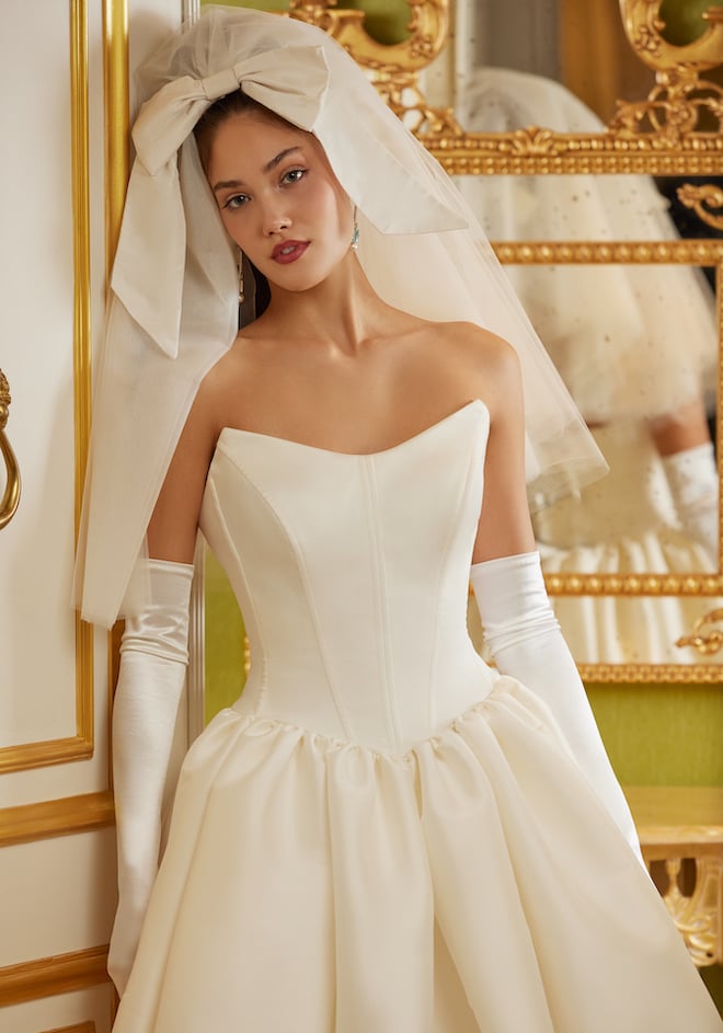 Model wearing a shoulder-length veil with a bow on the top and a strapless basque waist dress with satin gloves. 