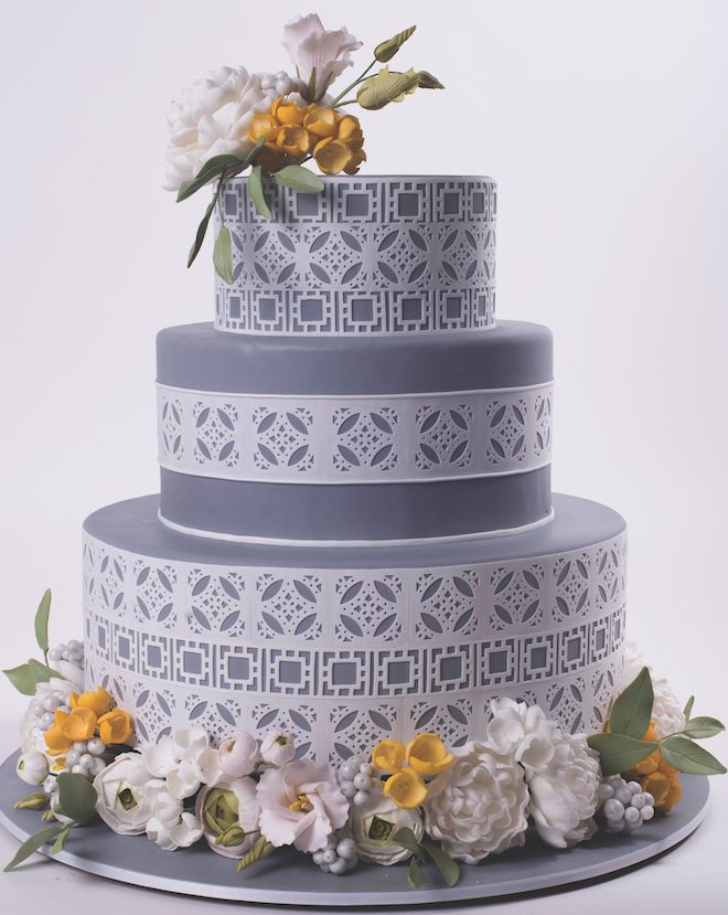 A three-tiered blue wedding cake is decorated in a white textured pattern with yellow and white flowers. 