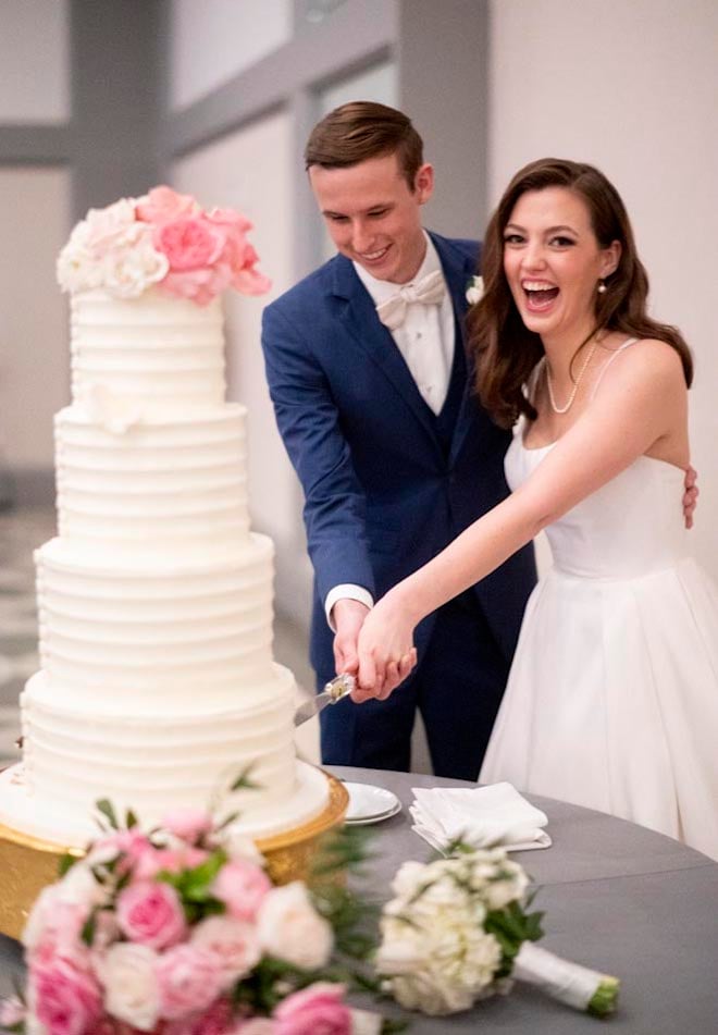 The bride and groom slice into their four-tier white wedding cake topped with pink roses. 