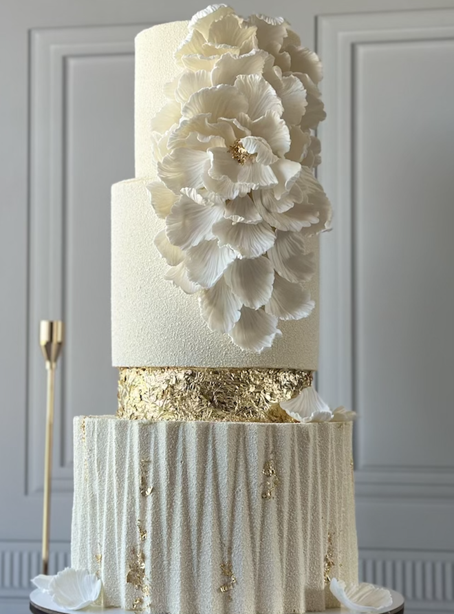 Romana Cakes designs a three-tiered textured white wedding cake with flowers and gold accents. 
