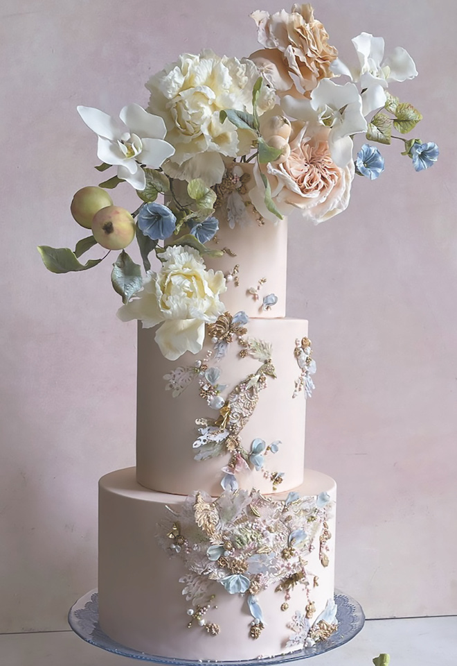 A pink wedding cake is topped with pastel florals and gold and light blue accents. 