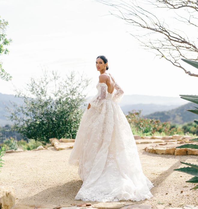 A bride wearing a lacy off the shoulder wedding gown by Monique Lhuillier in Ojai, California with the mountains behind her. 