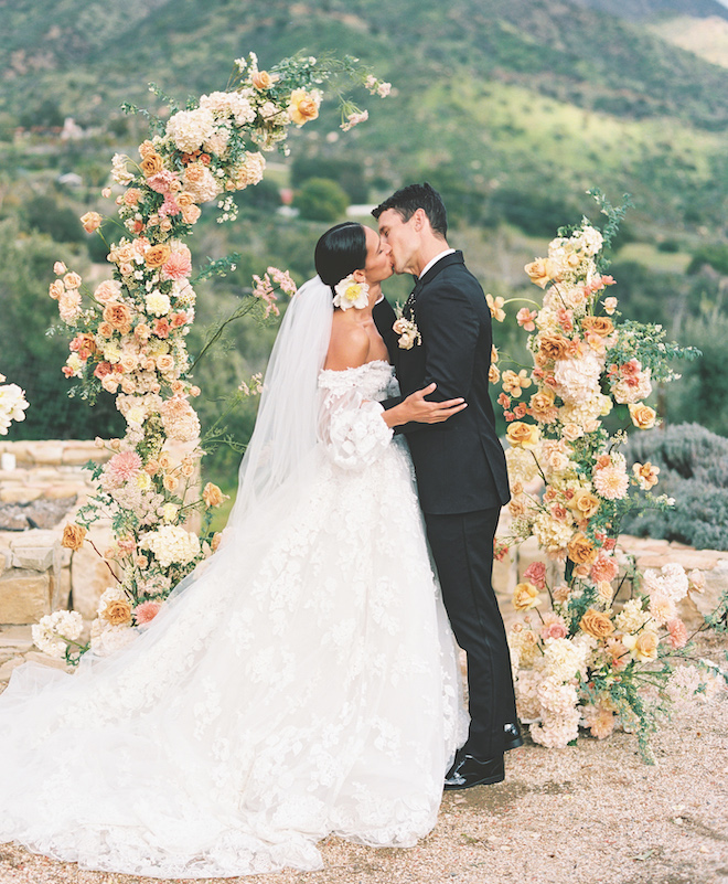 A bride and groom kissing in front of a peach-hue floral arrangement with the Ojai mountains behind them. 