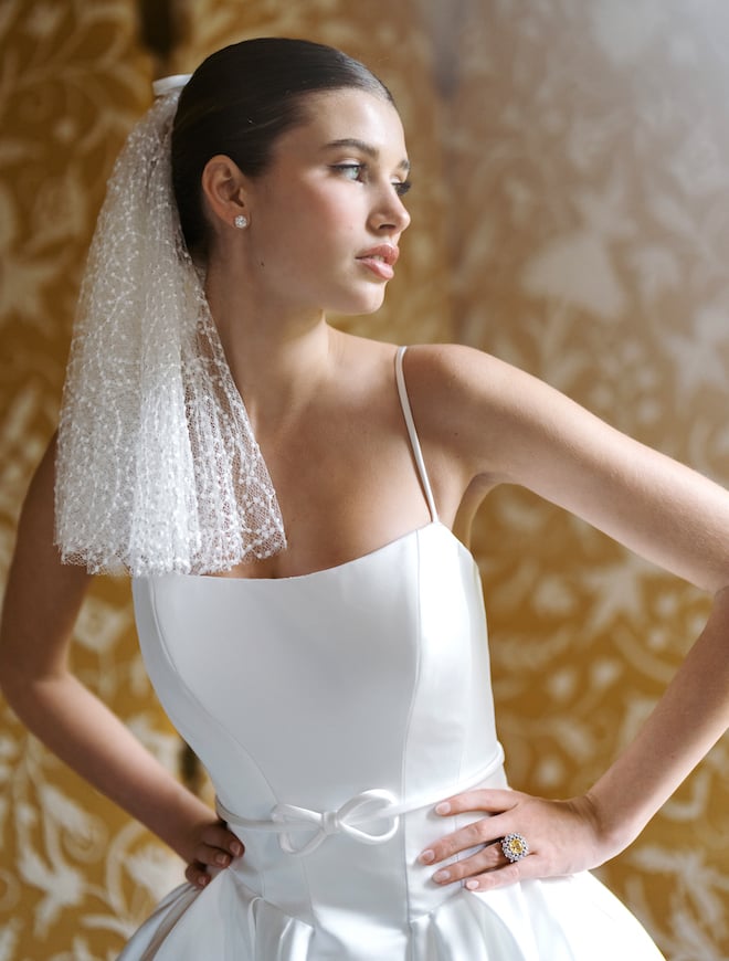 Model wearing a spaghetti stap wedding gown with a bow at the waist and a shoulder-length detailed veil. 