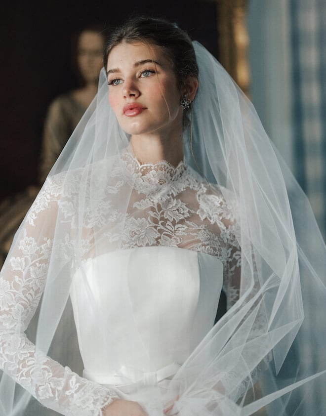 Model wearing a wedding gown with a high-neck long sleeve lace overlay and a sheer tulle veil. 