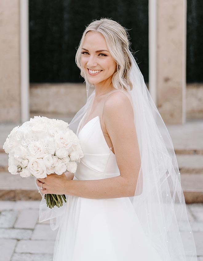 Bride smiling while wearing her wedding gown and veil and holding a bouquet of all-white flowers. 