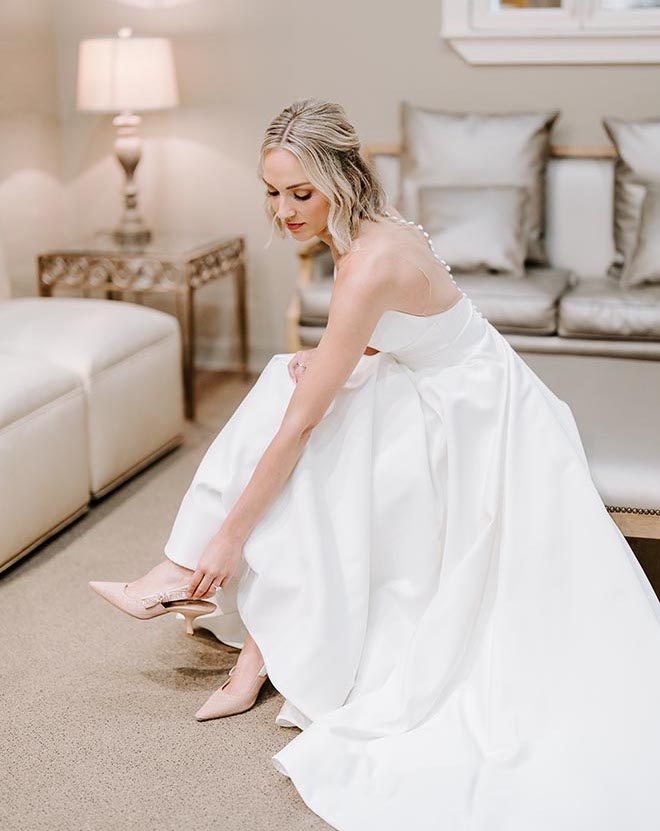 A bride wearing a wedding dress putting on a pair of nude pointed toe heels. 