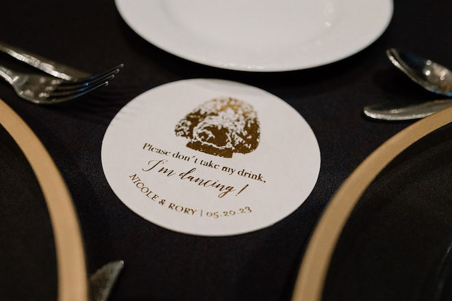 A white coaster on a black table with a gold etched photo of a dog with words underneath reading "Please don't take my drink, I'm Dancing" with the couples' name and wedding date.