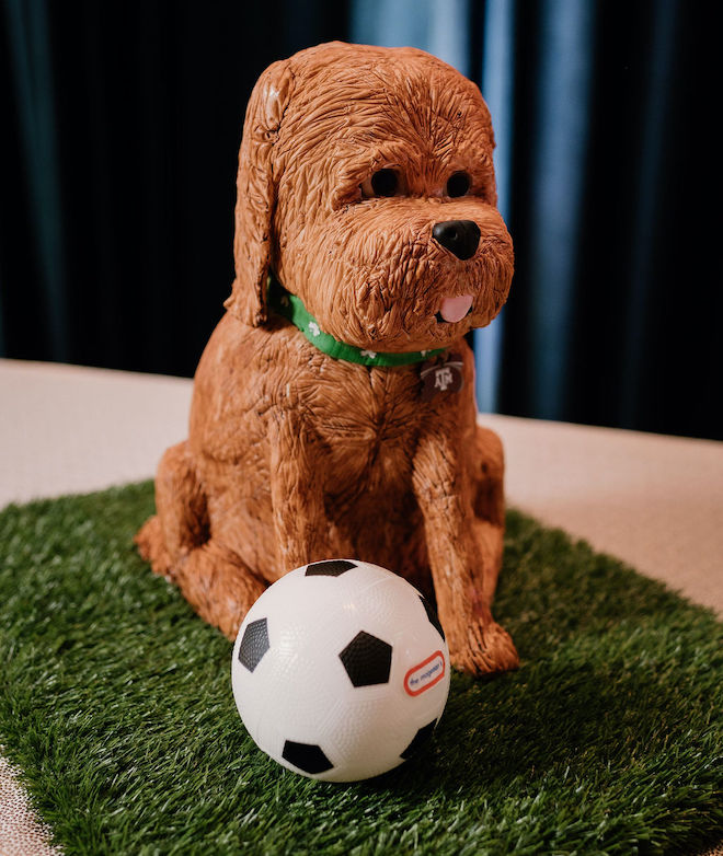 A groom's cake made to be a replica of a brown dog with it's tongue sticking out sitting on a patch of grass next to a soccer ball. 