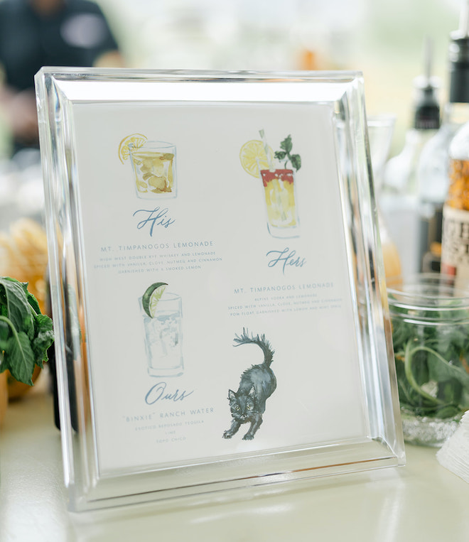 A silver framed cocktail menu with a his, hers and ours signature cocktail with a black cat in the corner of the menu. 