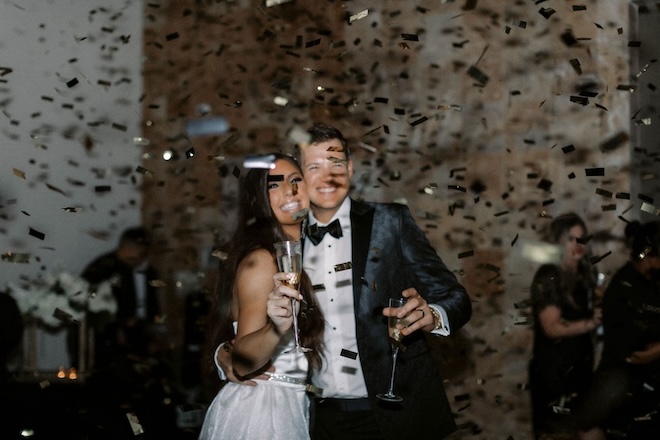 A bride and groom smiling while holding champagne with confetti raining on them.