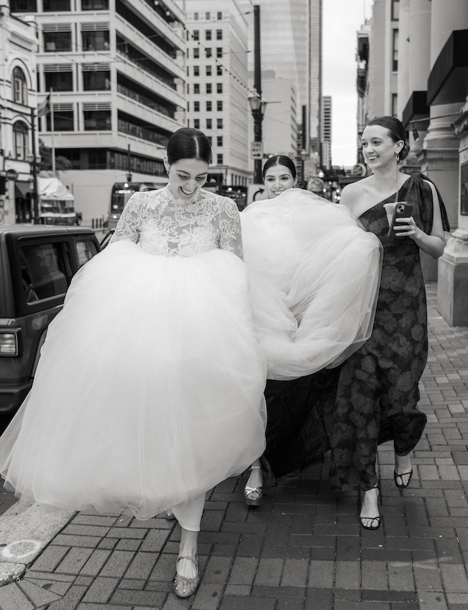 The bride smiling walking downtown with two bridesmaids holding up her gown.