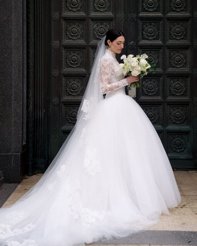 The bride holding a bouquet of white florals and wearing a wedding gown with a full tulle skirt and long lace sleeves.