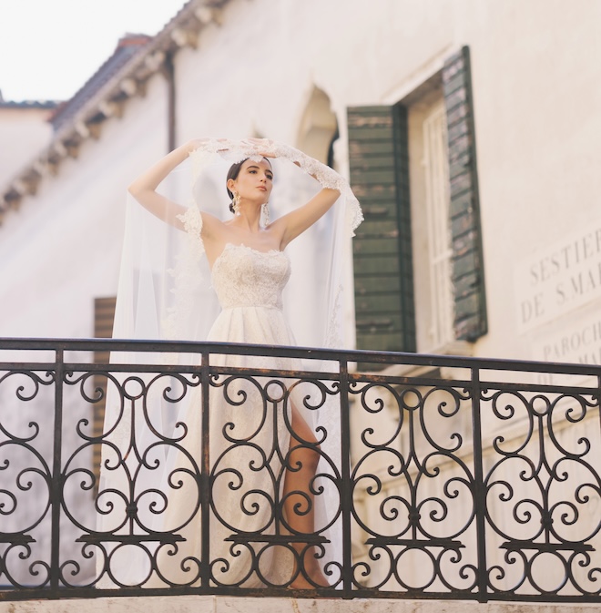 The bride standing on a balcony with her hands over her head while the veil drapes over her shoulders.