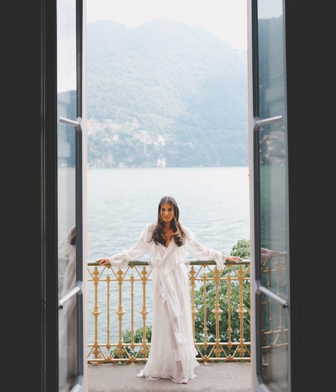 A bride standing on a balcony in front of Lake Como wearing a white ruffle robe.