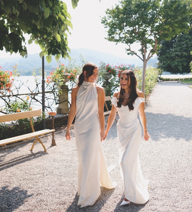 Two brides holding hands in wedding gowns overlooking the Lake Como scenery.