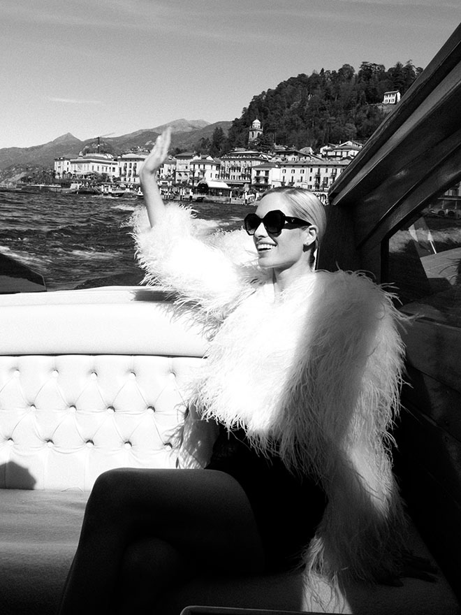 The bride wears a white furry jacket as she waves and rides a boat on Lake Como.