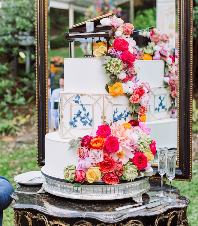 Three-tier wedding cake with a blue and gold design, a piano as the cake topper and bright florals garnished on the side. 