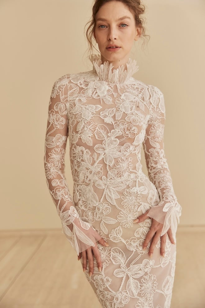 A long-sleeve gown featuring a high neckline with a dramatic ruffled collar and a slim, sculpted silhouette that flows into a soft, delicate train.