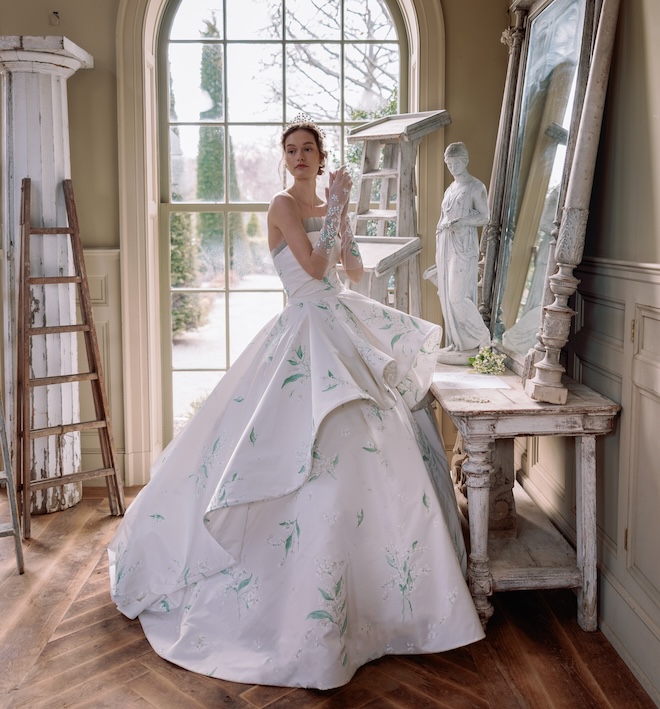 Lily of the Valley Radzimir gown, featuring a straight neckline with gathered tulle detail, a hand- draped bodice, and a dramatic layered oversized ballgown skirt with a cathedral-length train.
