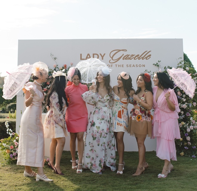 The bride and her friends standing in front of a white sign that says "Lady Gazelle Diamond of the Season" surrounded by florals.