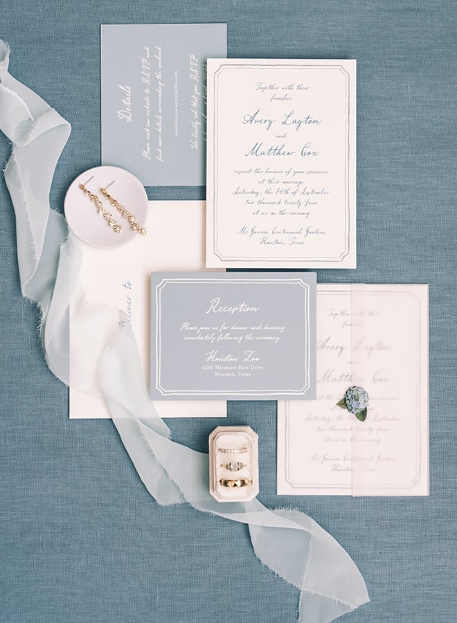 Blue and white wedding invitations and stationery with blue ribbon lay on a blue backdrop. 