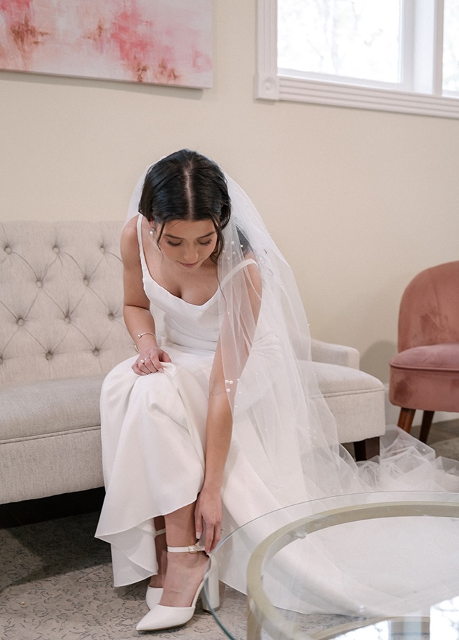 The bride fixes the straps on her wedding shoes while sitting on a beige couch in the bridal suite.