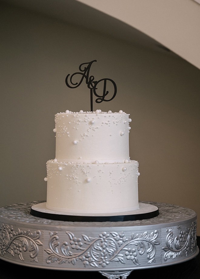 A white wedding cake embellished with pearls features a black cake topper with the bride and grooms initials. 