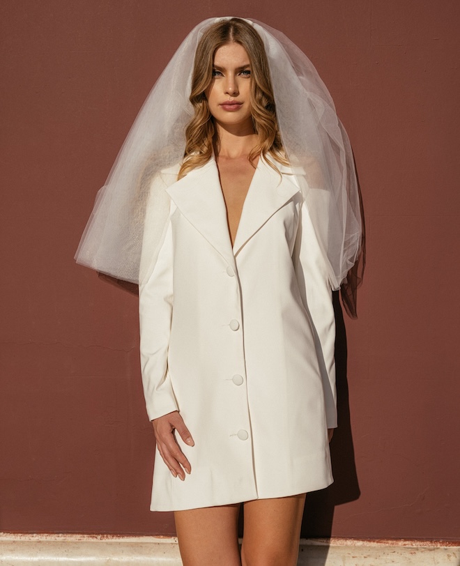 The bride wearing a short "blazer-style" dress and a veil in front of a maroon wall. 