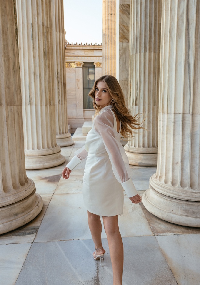 The bride looking over her shoulder walking in between pillars wearing a long-sleeve short dress at golden hour.