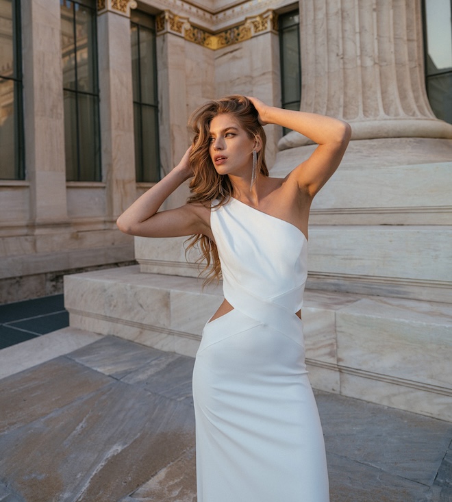 The bride wearing a one-shoulder wedding gown with her hands in her hair. 