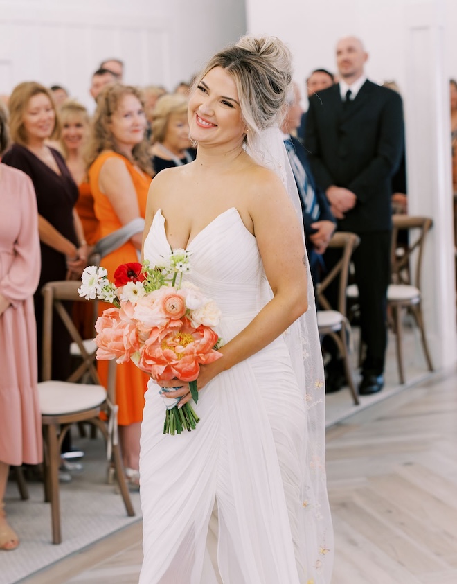 The bride smiling and holding pink and orange bouquet walking down the aisle. 