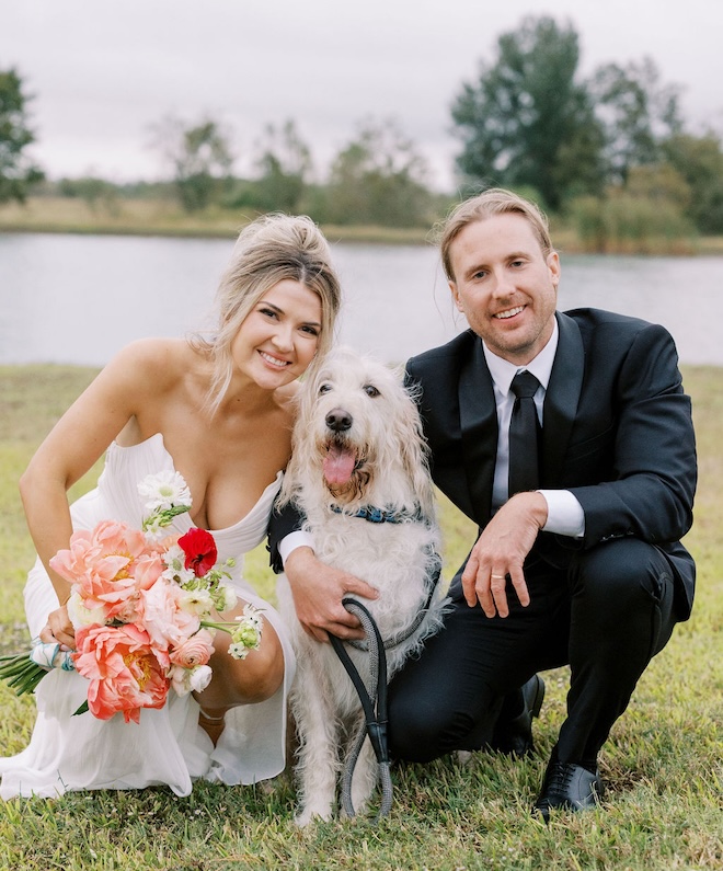 The bride and groom smiling with their dog in front of a pond. 