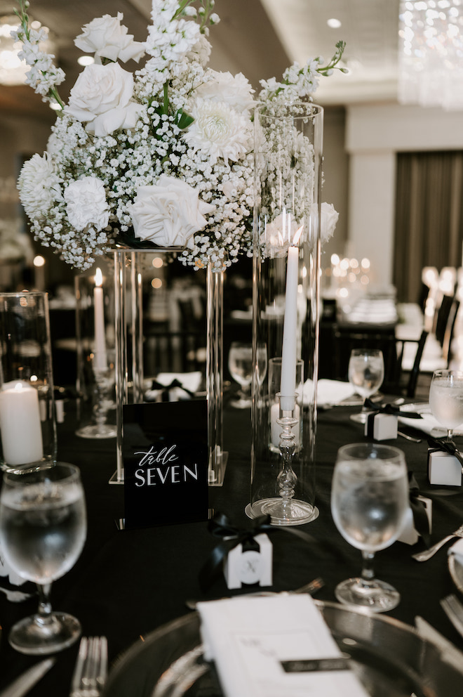 Table 7 settign at reception, black linen table cloth, white roses as the centerpiece and white candle sticls surrounding the centerpiece, each in a glass.