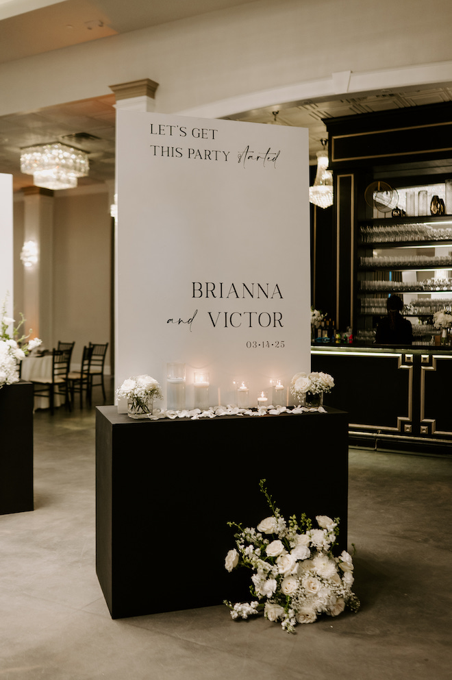 The white sign entering the reception that reads, "Let's get this party started, Brianna and Victor 3.14.25" on a black table surrounded by white florals and candles. 