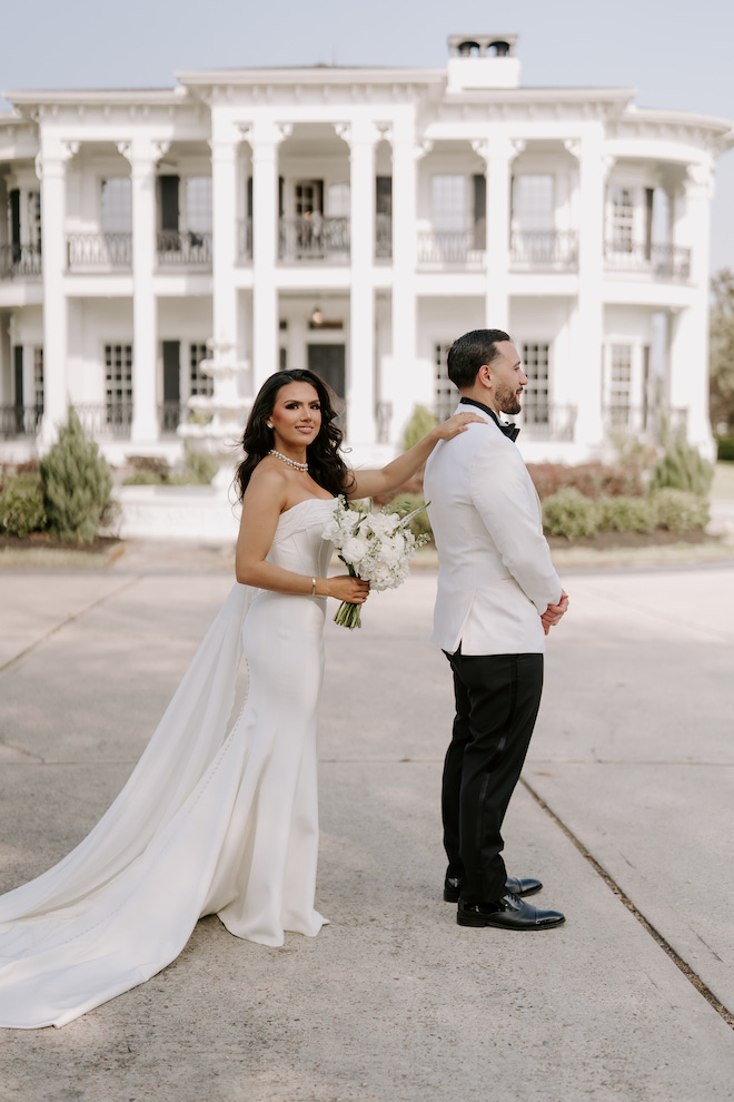 The bride, wearing a strapless wedding gown, tapping on the groom's shoulder while he is turned away for their first look outside of the venue.