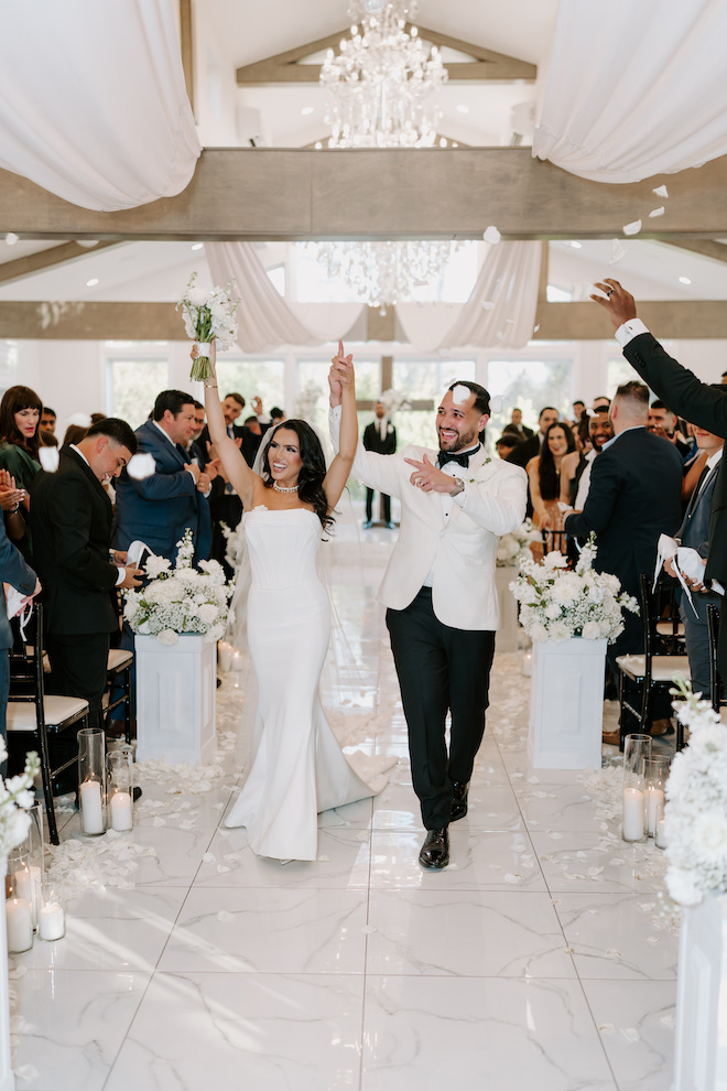 The couple holding hands and raising their arms smiling while the guests cheer as they walk down the aisle away from the altar. 