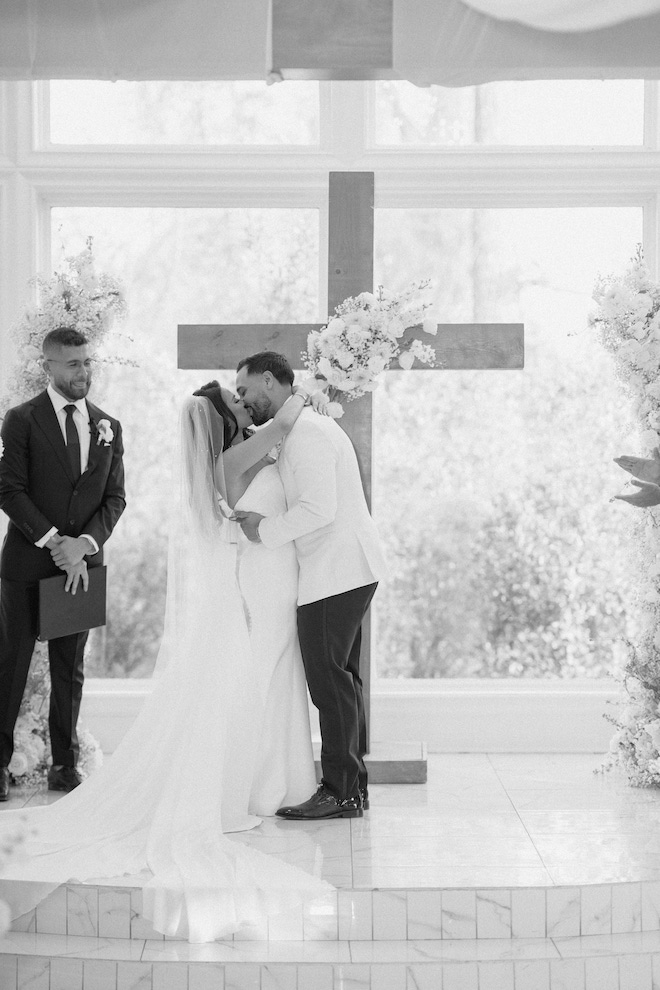 The couple kissing at the altar in front of a floral adorned cross in a black and white photo