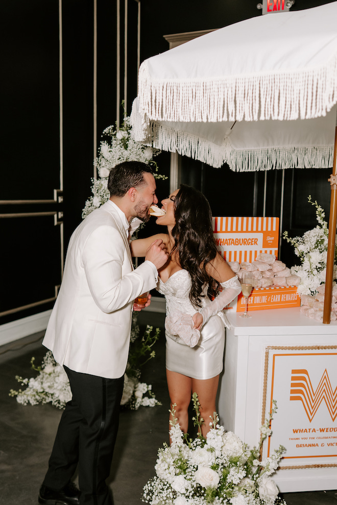 Bride, wearing her mom's wedding gown transformed into a modern mini, and groom biting into the same whataburger sandwhich next to their whataburger cart for the reception 