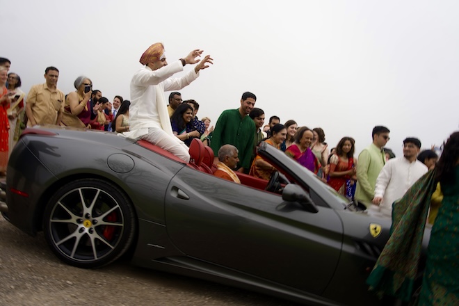 The groom rides sitting on the back of a black Porsche as family and friends watch. 
