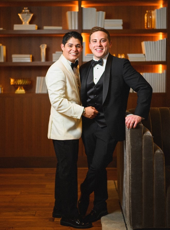Two grooms smiling on their wedding day in front of a bookshelf at Thompson Houston, by Hyatt. 