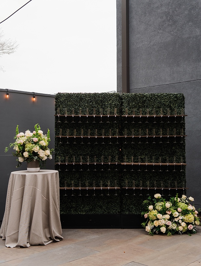 A champagne wall covered in greenery holds Champagne flutes for cocktail hour. 