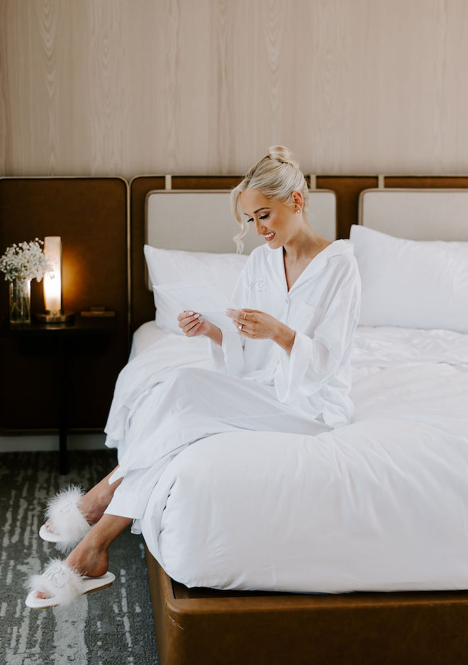 Bride sitting on hotel bed in silk pajamas reading her husbands pre wedding note.