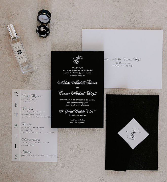 The black and white wedding invitations laying on the white tile floor with the ring and her perfume laying above them.