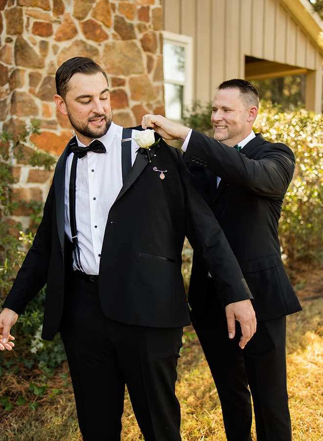 A groomsmen helps the groom put on his tuxedo jacket. 