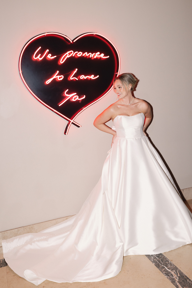 Bride smiling and standing next to pink heart sign that reads, "We promise to love you".