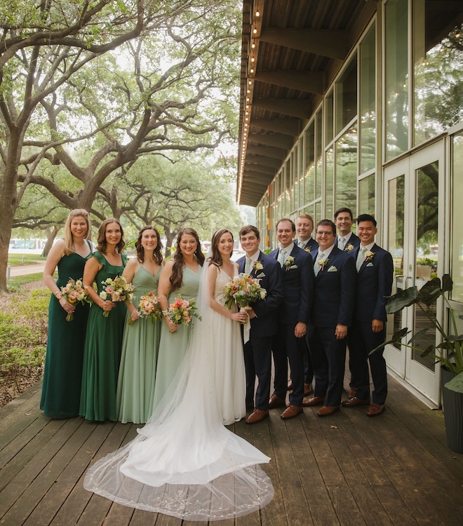 The bride and groom smiling with their wedding party outside The Grove. The men are wearing blue suits and the bridesmaids are wearing green dresses. 