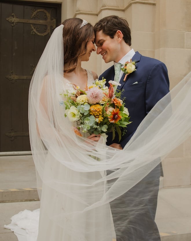 The bride and groom smiling outside the church while touching foreheads while the bride's veil flowed. 